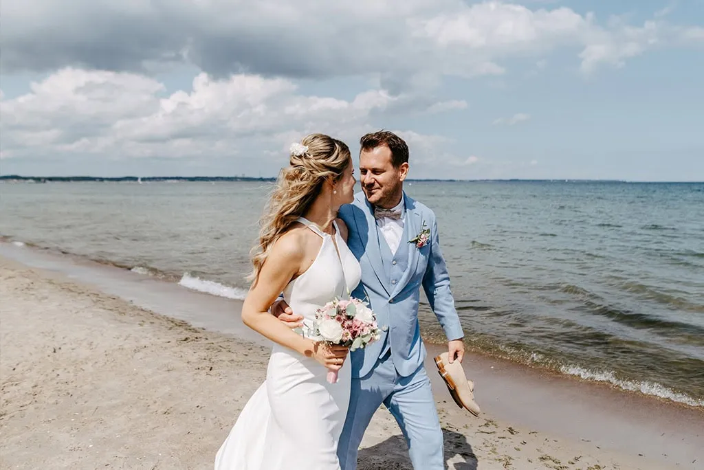 Braut und Bräutigam spazieren am Sandstrand entlang, halten sich an den Händen und lächeln sich an, mit dem Meer und dem wolkenverhangenen Himmel im Hintergrund - wunderschön fotografiert von Hochzeitsfotograf Ostsee Stefanie Roth Fotografie.